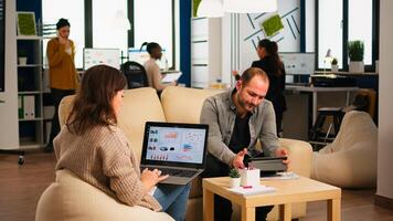 Back view of young business woman sitting on couch in office using laptop computer while colleagues working on background. Multiethnic coworkers planning new financial project in modern company photo