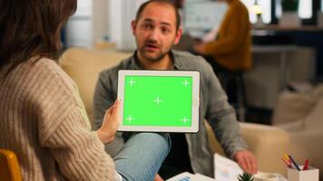 Over the sholder of manager woman sitting on couch talking with coworker holding tablet with green screen while diverse team working. Multiethnic people planning project on chroma key display photo