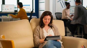 Employee taking break sitting on confortable couch holding tablet browsing, reading great news and smiling. Multiethnic creative coworkers planning new financial project working in open plan office photo