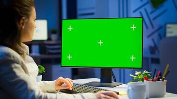 Freelancer working in front of green screen display sitting at desk in business office late at night. Businesswoman watching desktop laptop monitor with green mockup, chroma key, working overtime. photo