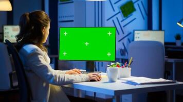 Businesswoman looking at green screen monitor of computer sitting at desk in business office late at night. Freelancer watching desktop monitor display with green mockup, chroma key working overtime. photo