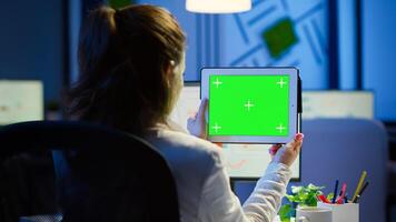 Businesswoman holding tablet with green screen monitor sitting at desk in business office late at night. Freelancer watching desktop monitor display with green mockup, chroma key working overtime. photo