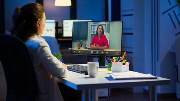 Freelancer working remotely discussing with woman partner online sitting in front of computer in start-up office talking during virtual meeting at midnight, using wireless headphone doing overtime photo