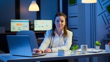 Business woman looking at camera smiling after typing on laptop sitting at desk in start-up company late at night. Focused employee using technology network wireless doing overtime for job reading. photo