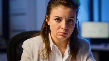 Close up of overworked business woman looking exhausted at camera sighing working late at night in start-up business office. Focused employee using technology network wireless doing overtime for job photo