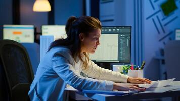 Focused woman architect checking and matching blueprints sitting at desk in front of computer late at night. Designer using arhitecture plans of buildings working overtime, creating and studying photo