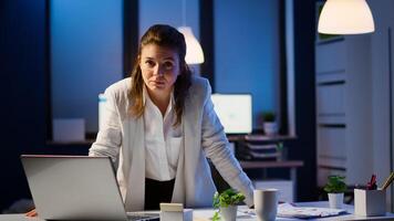 Business woman looking at camera smiling after typing on laptop standing near desk in start-up company late at night. Focused employee using technology network wireless doing overtime for job reading. photo