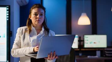 Tired manager raising head looking at camera sighing holding laptop typing on it, working late at night in start-up business office. Focused employee using technology network wireless doing overtime photo