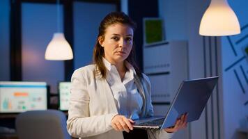 Exhausted business woman holding laptop, typing and sighing looking at camera standing in start-up company late at night. Employee using technology network wireless doing overtime for job reading. photo