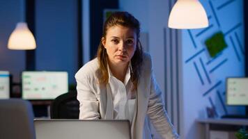 Focused tired business woman looking at camera after reading mails on laptop standing near desk in start-up company late at night. Focused employee using technology network wireless doing overtime photo