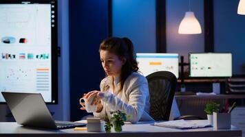 Business woman working overtime checking team project, taking notes, drinking coffee, analysing financial documents sitting at desk in start-up company late at night using modern technology network photo