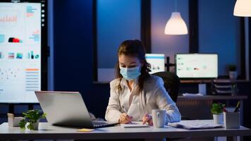Manager woman with face mask working overtime in new normal business office checking team project, taking notes, analysing financial documents sitting at desk late at night during global pandemic photo