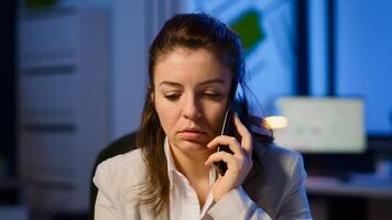 Tired freelancer woman having conversation on phone while working exhausted in business office late at night doing overtime. Focused employee using modern technology network wireless overworking photo