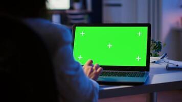 Close up of freelancer typing on laptop with green screen sitting at desk in start up office overworking. Manager watching desktop monitor display with green mockup, chroma key, during night time photo