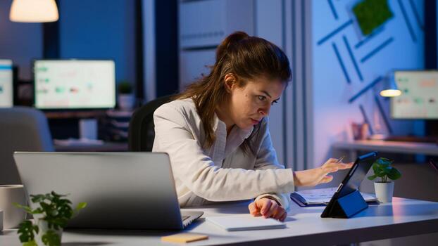 Manager woman using laptop and tablet in same time working overtime in business start-up office. Busy multitasking employee using modern technology network wireless overworking writing, searching. photo