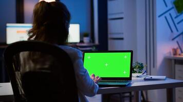 Back view of business woman looking at green screen laptop sitting at desk in start up office overworking. Freelancer watching desktop monitor display with green mockup, chroma key, during night time photo