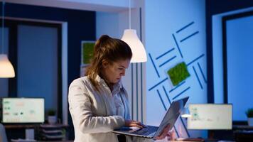 Busy entrepreneur analysing financial reports standing in start up office late at night holding laptop. Focused employee using technology network wireless doing overtime reading writing, searching photo