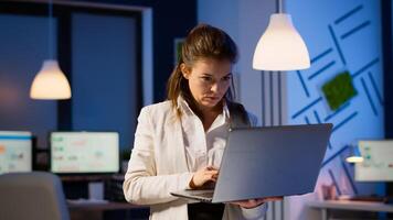 Focused businesswoman checking business reports standing in start up office late at night typing on laptop. Busy employee using technology network wireless doing overtime reading writing, searching photo
