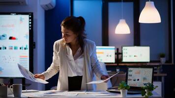 Stressed manager woman working with financial documents checking graphs, holding papers, reading raports, standing at desk late at night in start-up office doing overtime to respect deadline. photo