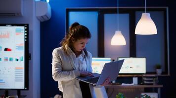 Focused manager woman checking business reports standing in start up office late at night holding laptop. Busy employee using technology network wireless doing overtime reading writing, searching photo