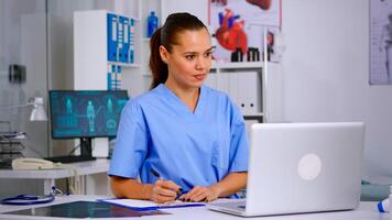 Doctor assistant in uniform working on laptop in modern hospital office. Therapist writing list of consulted patients online, making research, noting, taking useful information on clipboard photo