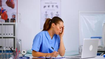Tired healthcare medical assistant working in hospital researching diagnosing patients health using laptop taking off the glasses having headache. Exhausted therapist in medicine uniform resting eyes photo