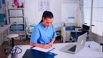 Healthcare assistant taking notes on clipboard and typing on laptop working in hospital clinic. Medical physician in medicine uniform writing list of consulted, diagnosed patients, making research. photo
