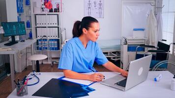 Therapist doctor assistant in uniform writing on clipboard, checking list of patients working on laptop in hospital clinic. Noting consulted patients online, making research, useful information photo