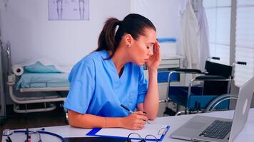 Overworked nurse wrinting on clipboard, working on laptop, taking off the glasses relaxing eyes. Medical physician in medicine uniform writing list of consulted, diagnosed patients, making research. photo