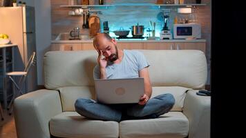Tired man in pajams browsing on internet lifestyle information using laptop computer sitting on sofa. Freelancer working at online project in front on television late at night in kitchen photo