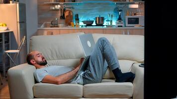 Exhausted man falling asleep while working on internet communication project using laptop computer with modern technology wireless. Caucasian male in pajamas sitting on sofa late at night in kitchen photo