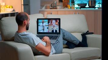 Comfortable man relaxing on couch while having online business conference with collegues using laptop computer during quarantine. Focused adult talking on videocall use webcam late at night in kitchen photo