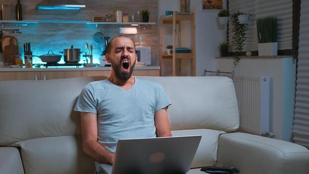 Focused man browsing information on network using laptop computer sitting in front of television. Caucaisan male lying on sofa late at night in kitchen using modern technology wireless photo