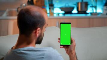Caucasian male reading online social information on phone with mock up green screen chroma key display. Tired man sitting on sofa using isolated smartphone with technology internet photo