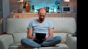 Tired man sitting alone on couch while browsing on internet using tablet computer with modern technology wireless. Caucasian male in pajamas relaxing in front of televion late at night in kitchen photo