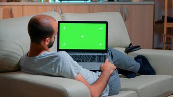 Tired man lying on sofa working on computer with mock up green screen chroma key display searching online information. Caucasian male using isolated pc late at night in kitchen photo
