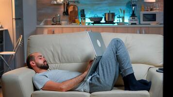 Man falling asleep in fron of TV while working on the laptop. Cozy man lying down on sofa while typing on laptop computer for social media project. Exhausted man working photo