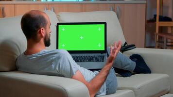 Focused man looking laptop computer with at mock up green screen chroma key display while laying on couch working at online e-learning project. Tired person using isolated pc late at night in kitchen photo