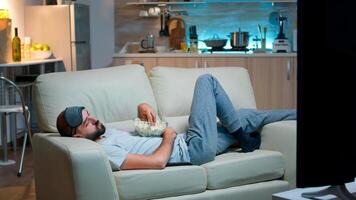 Man lying on sofa, falling asleep in fron of the TV with popcorn bowl in his hands. Tired alone man in living room after a hard day at work photo