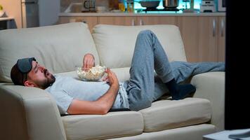 Caucasian man lying in sofa, eating popcron and watching TV late at night in the living room. Enjoying midnight snack, television entertainment, leisure lifestyle evening show photo