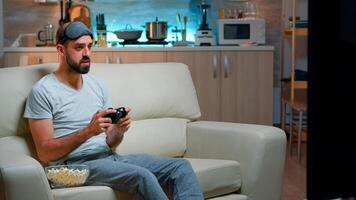 Frustrated pro gamer losing entertainment gaming competition using joystick. Serious man sitting on couch late at night in kitchen while playing online games on television photo