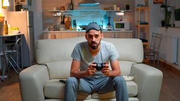 Focused man holding joystick while sitting in front of television winning online games competition. Caucasian male with sleep mask lying on couch making winner gesture late at night in kitchen photo