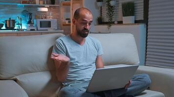 Focused man sitting on sofa in pajamas while talking with collegue during videocall conference using laptop computer. Caucasian male working at communication project late at night in kitchen photo