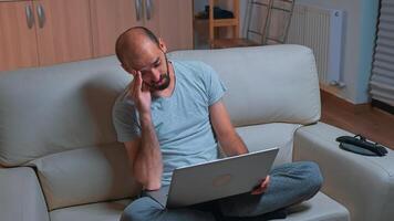 Tired man sitting on sofa while talking with collegue during business videocall conference using laptop computer, Caucasian male working from home because of coronavirus quarantine late at night photo