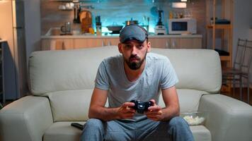 Front view of pro gamer sitting on sofa in front of television while playing videogames competition using wireless joystick. Cheerful man with sleep mask enjoying time alone late at night in kitchen photo