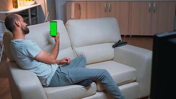 Caucasian male looking phone with at mock up green screen chroma key display while lying on sofa browsing on internet. Caucasian male using modern technology wireless late at night in kitchen photo