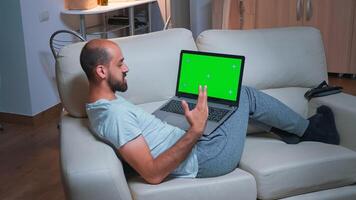 Tired man sitting on sofa while browsing marketing information using laptop computer with mock up green screen chroma key display. Caucasian male using modern technology wireless late at night photo