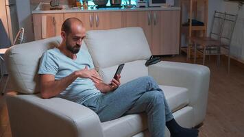 Focused adult talking with friends about social media connection using modern smartphone while sitting on couch. Tired man working late at night in kitchen using modern technology wireless photo