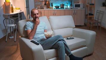 Man with beard watching tv show using remote control to change channels on television. Caucasian male relaxing on couch with popcorn bowl in hands looking entertainment series at late at night photo