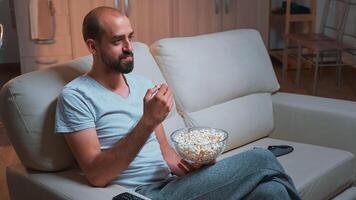 Caucasian male chilling on sofa with popcorn bowl in hands while watching movie series on television. Tired man in pajamas looking at entertainment shows late at night in kitchen photo